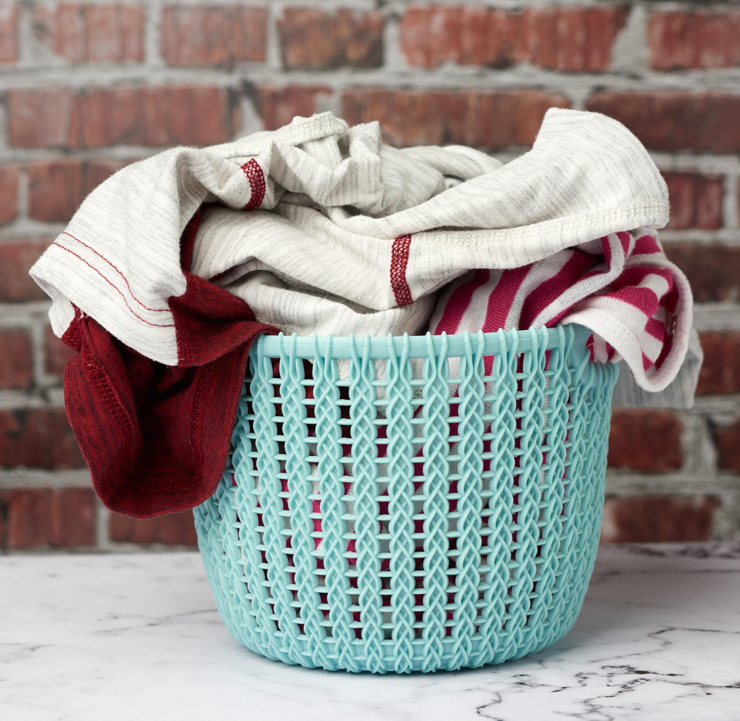 Teal laundry basket filled with clean red and white clothes against brick wall background, symbolizing Sudsies laundry by the pound service.