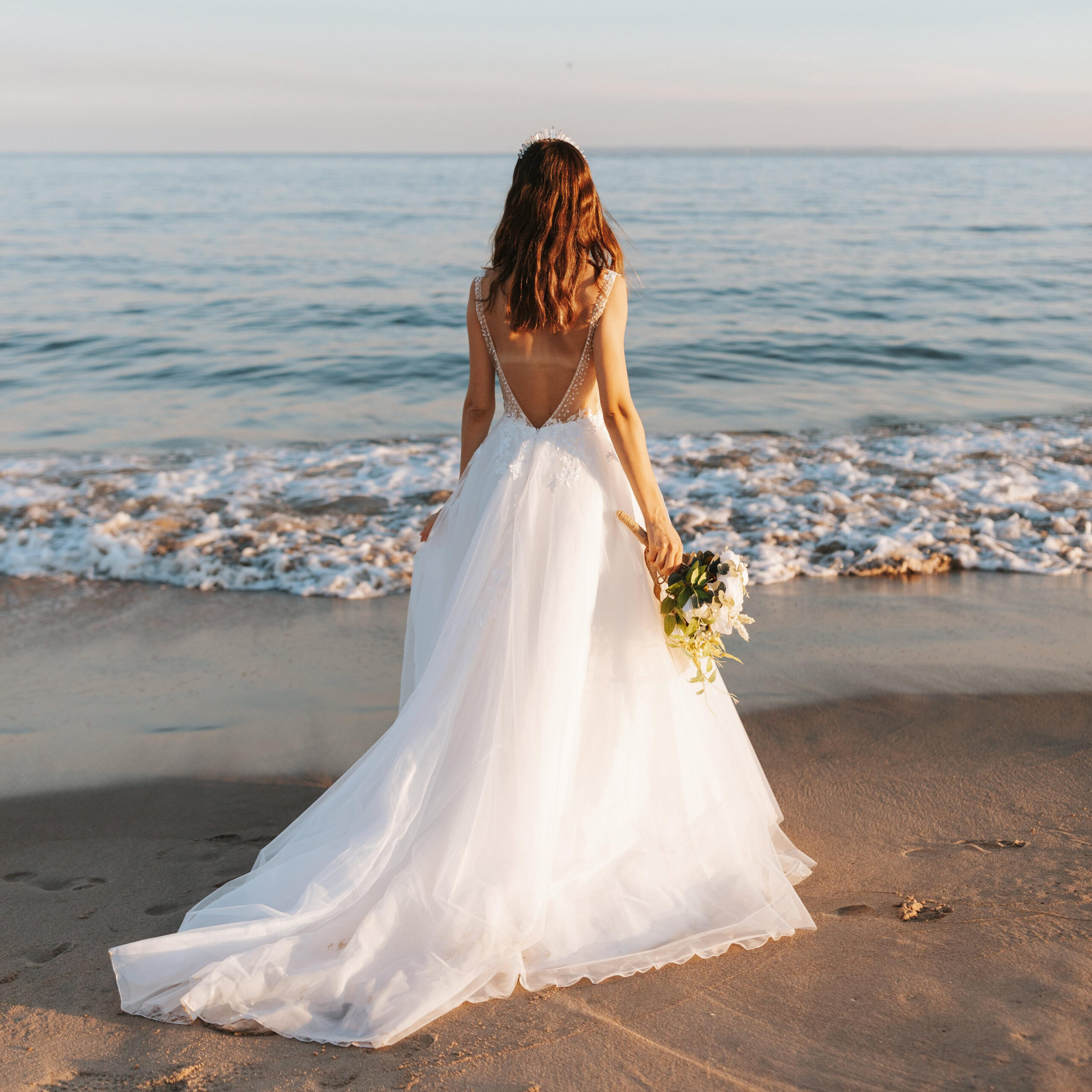 Bride in white wedding gown holding bouquet and standing on the beach facing the ocean at sunset, showcasing Sudsies bridal cleaning and preservation service.