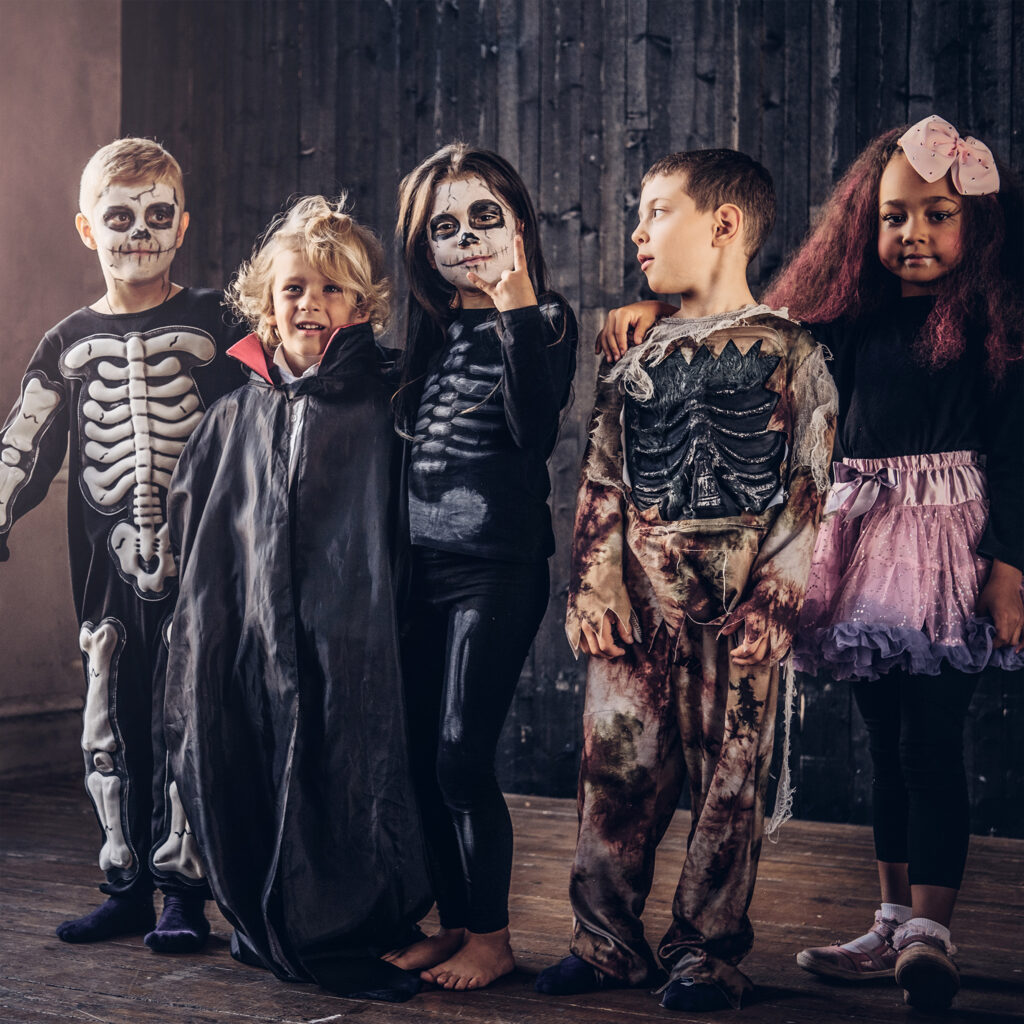 Group of children wearing Halloween costumes and face paint, standing together indoors and smiling.