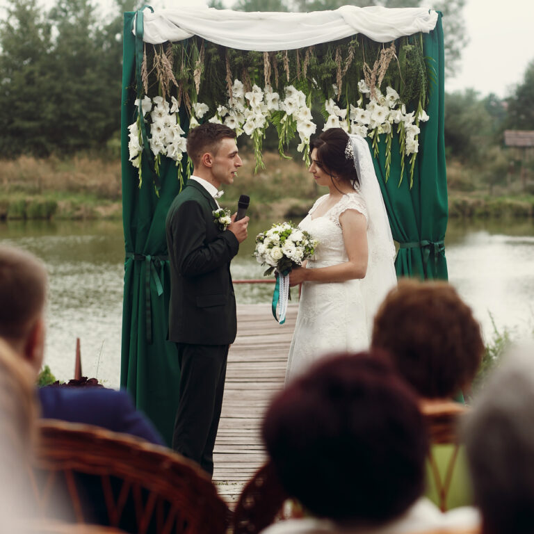 Bride and groom exchanging vows at an outdoor lakefront ceremony with floral arch, highlighting Sudsies’ bridal alterations and preservation services.