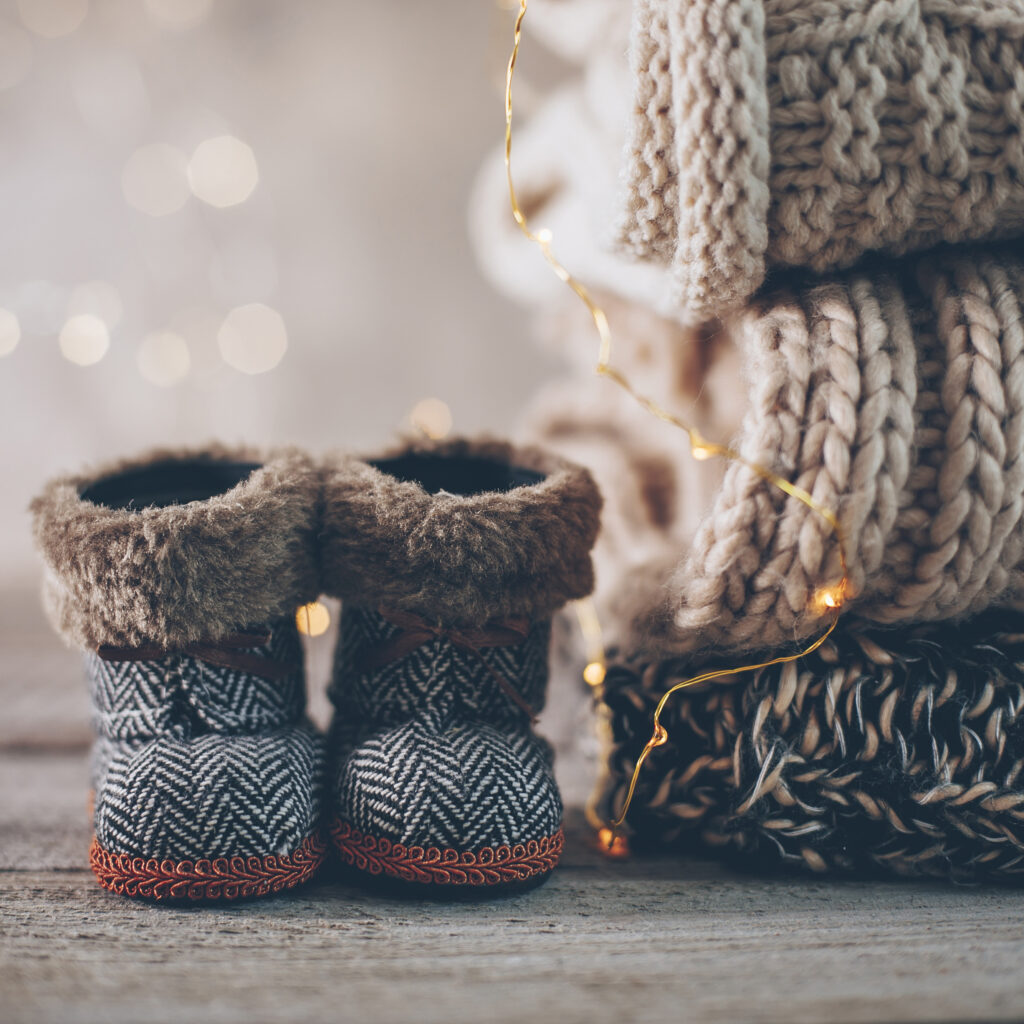 Stack of cozy knitted winter clothes and baby boots with warm lighting, illustrating cold weather garment care tips from Sudsies.