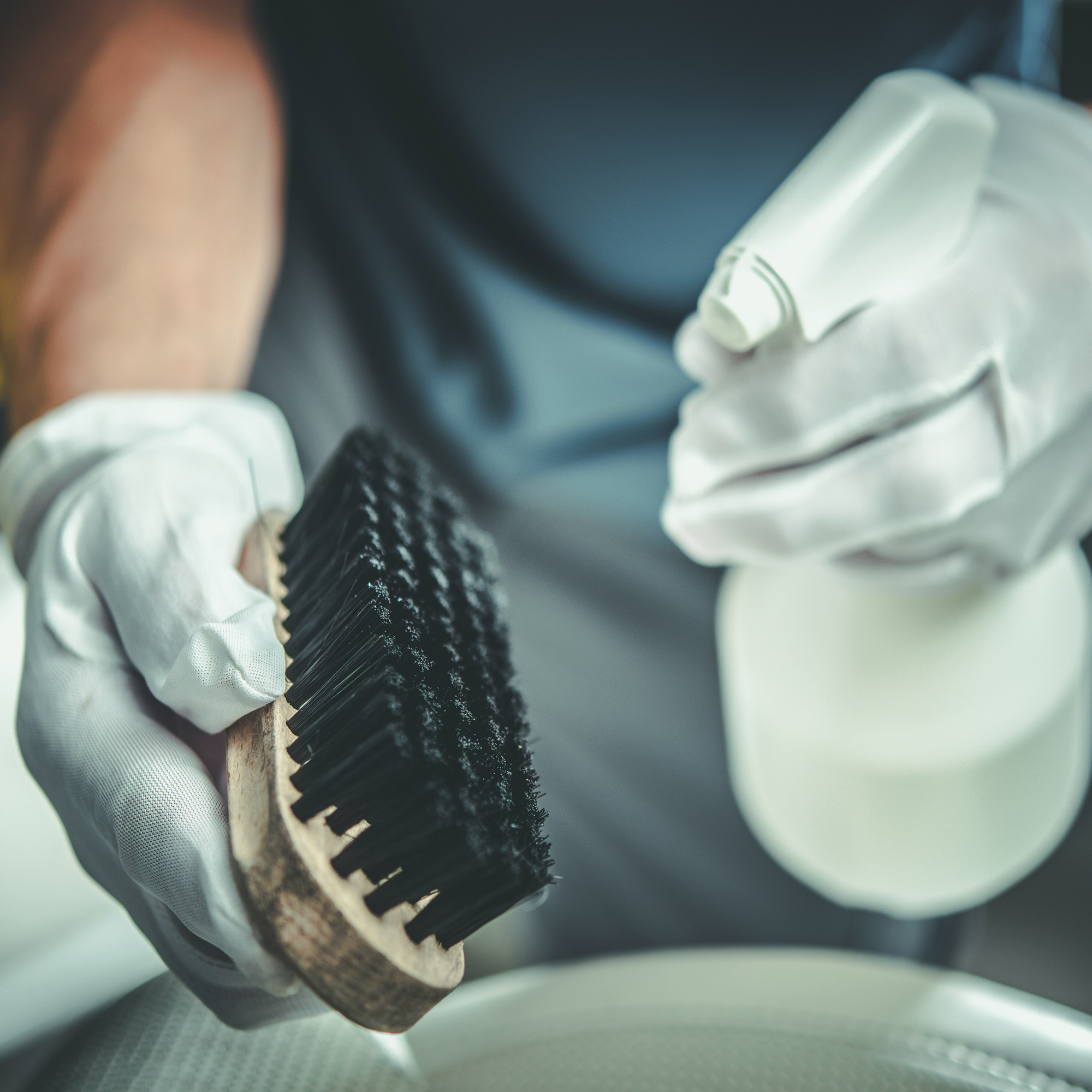 Gloved hands holding a garment cleaning brush and spray bottle during fabric treatment
