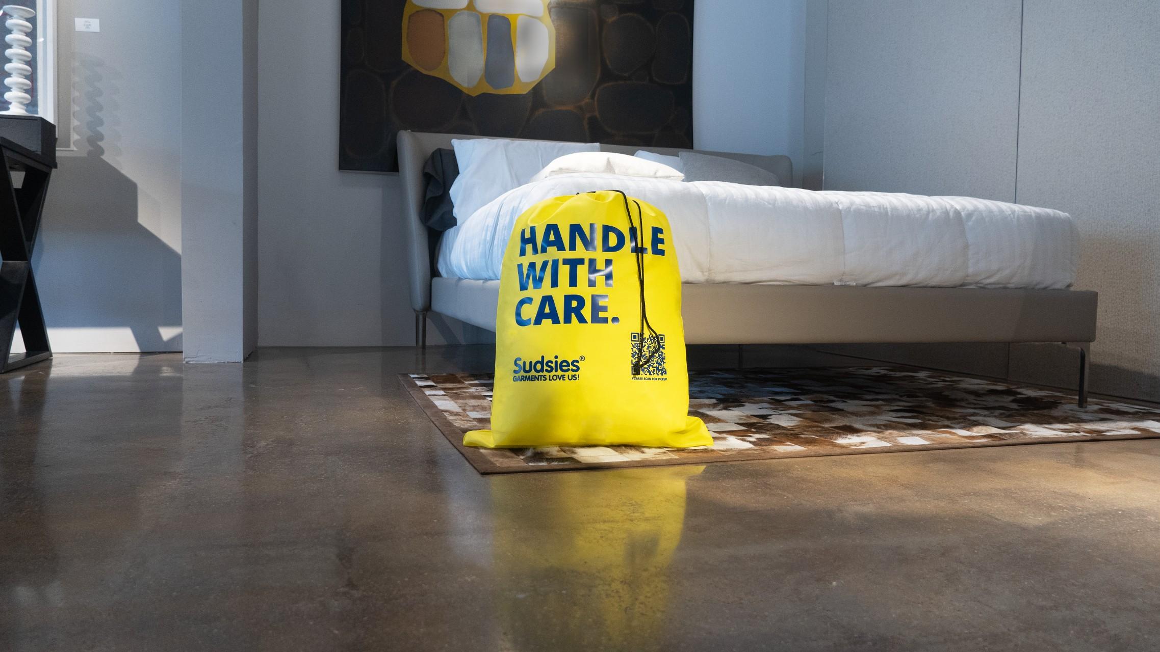 Yellow Sudsies laundry bag labeled "Handle With Care" placed in a modern bedroom beside a freshly made bed