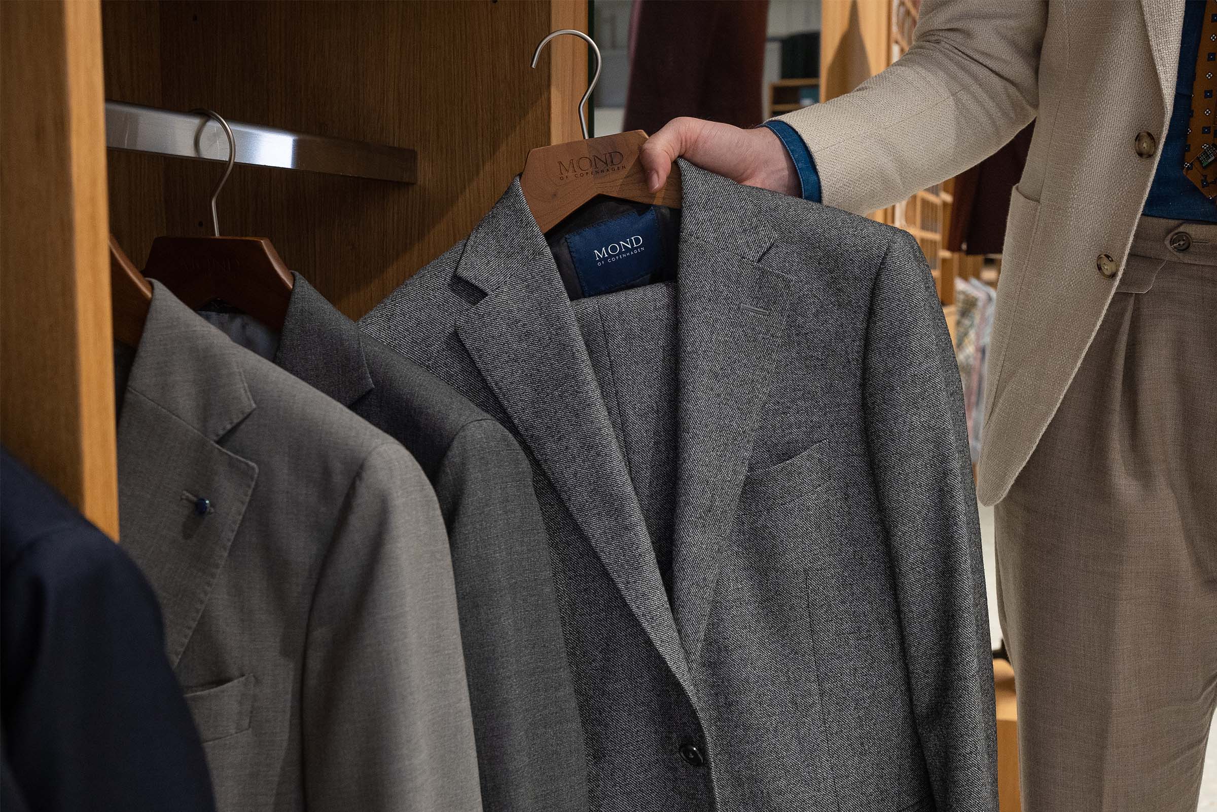 A person selects a Mond gray suit jacket from a refined wooden closet