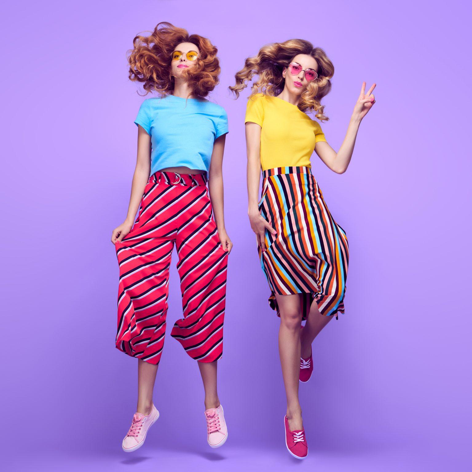 Two fashionable women in bright, striped outfits jumping against purple background