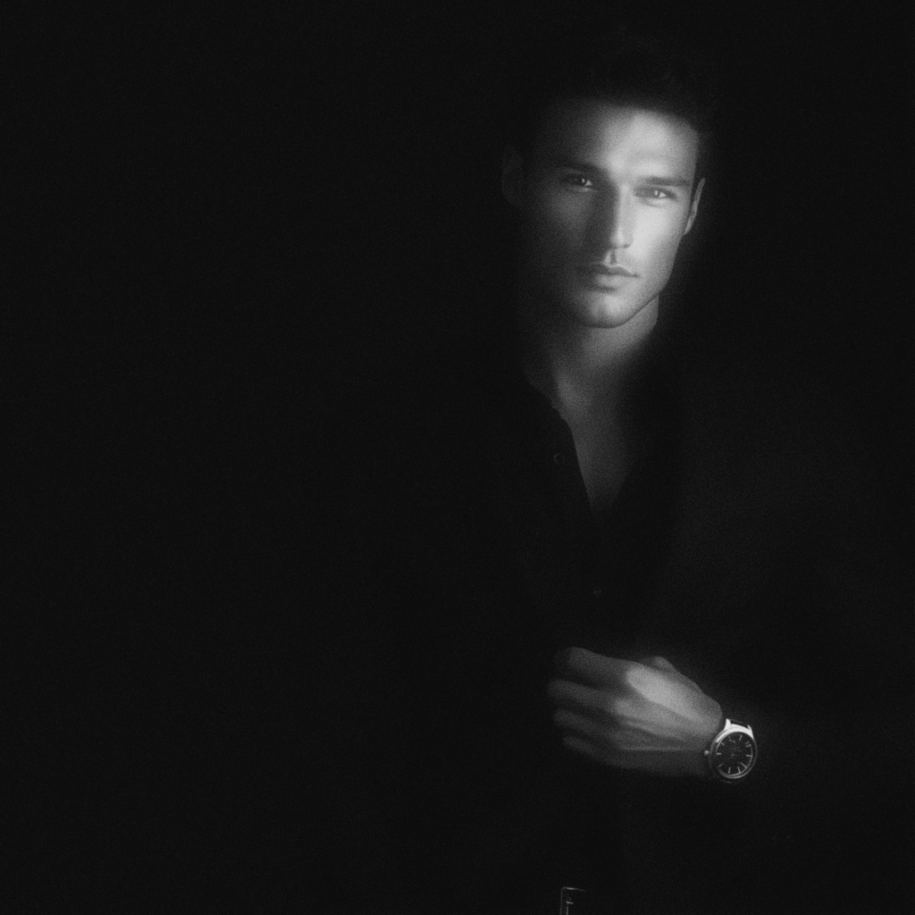 Black and white portrait of an elegant man in a dark tailored suit and watch against a black background, representing the quiet power of Kiton perfection.