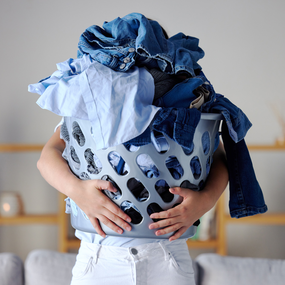 Person holding an overflowing laundry basket full of blue clothes, with their face hidden behind the pile.