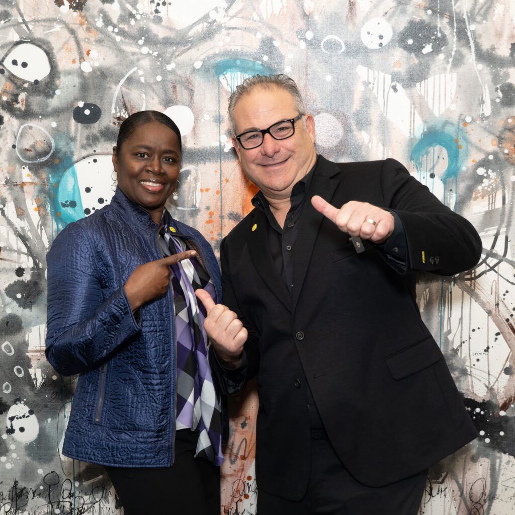 wo Sudsies team members posing in front of a colorful mural, smiling and pointing