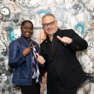 Sudsies Team Members at Event Photo Wall (Community & Culture) | Sudsies wo Sudsies team members posing in front of a colorful mural, smiling and pointing