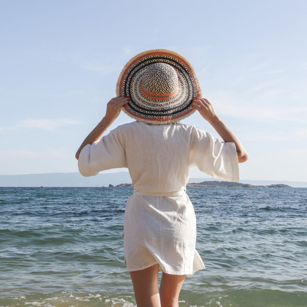 Woman in a white cover-up holding a wide-brim hat while standing at the ocean shore
