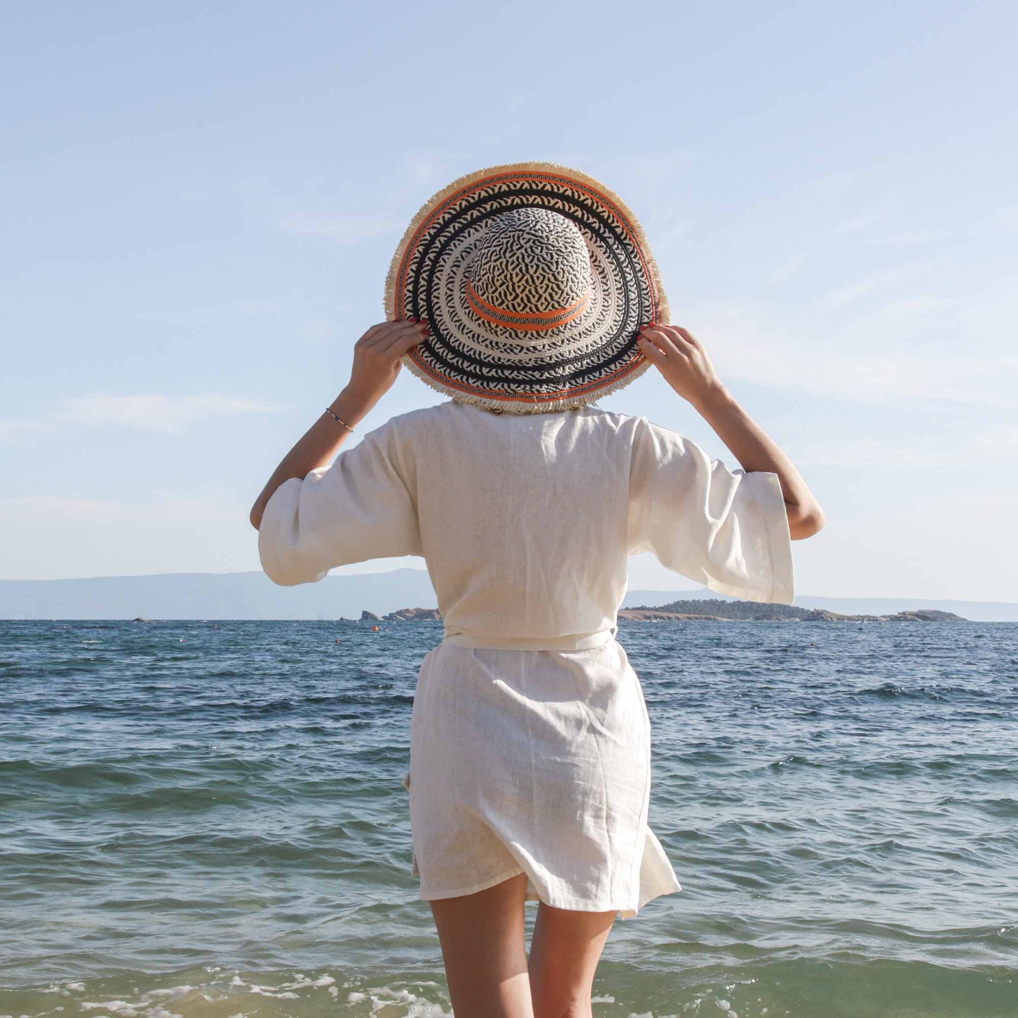 Woman in a white cover-up holding a wide-brim hat while standing at the ocean shore