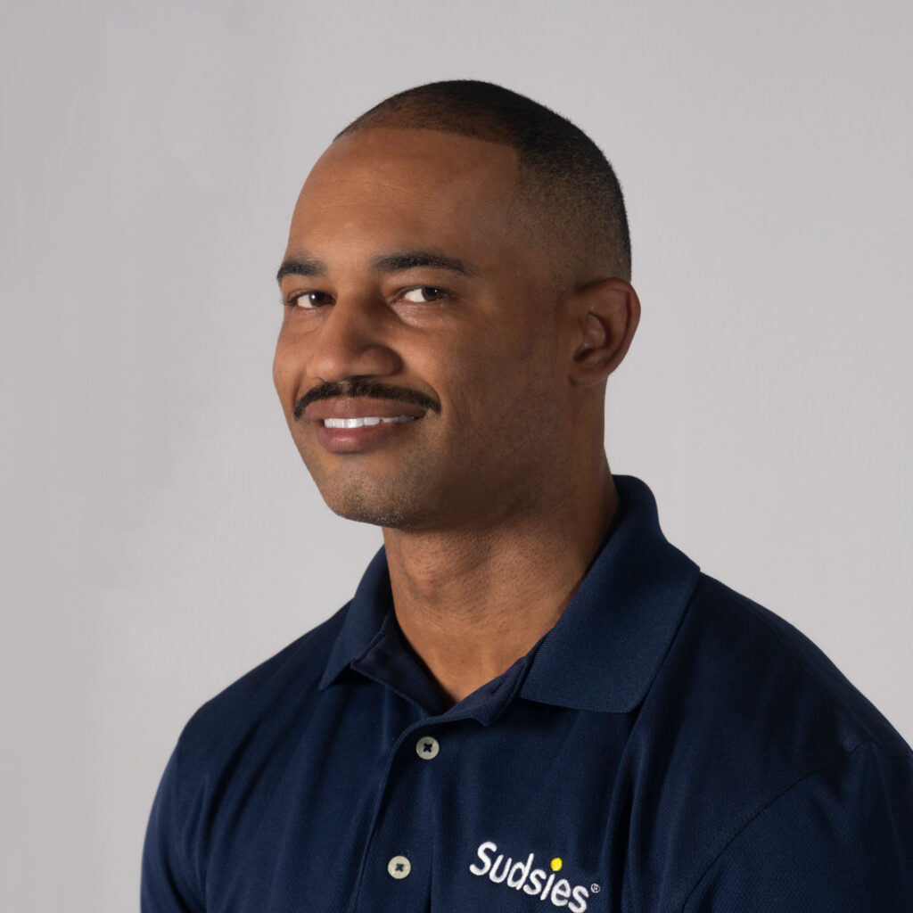 Portrait of a smiling man with short-cropped hair wearing a navy polo shirt with a Sudsies logo.