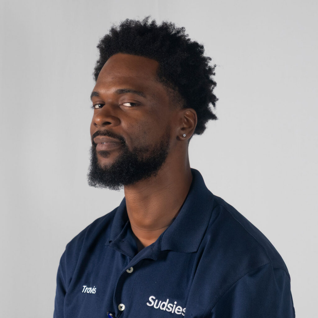 Portrait of Travis, a Sudsies employee, wearing a navy polo and a small earring, looking slightly to the left.