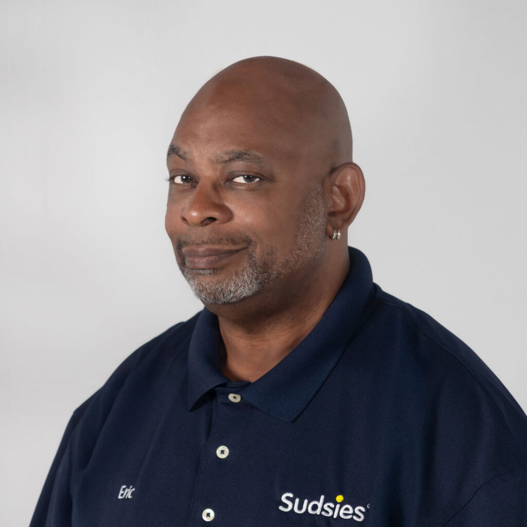 Portrait of a smiling man in a navy polo with Sudsies logo; name 'Eric' embroidered on chest, light gray background