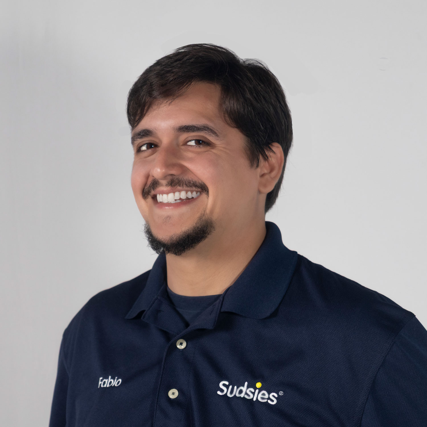 Head-and-shoulders portrait of a smiling man with dark hair and a goatee, wearing a navy polo labeled 'Fabio' and 'Sudsies'.