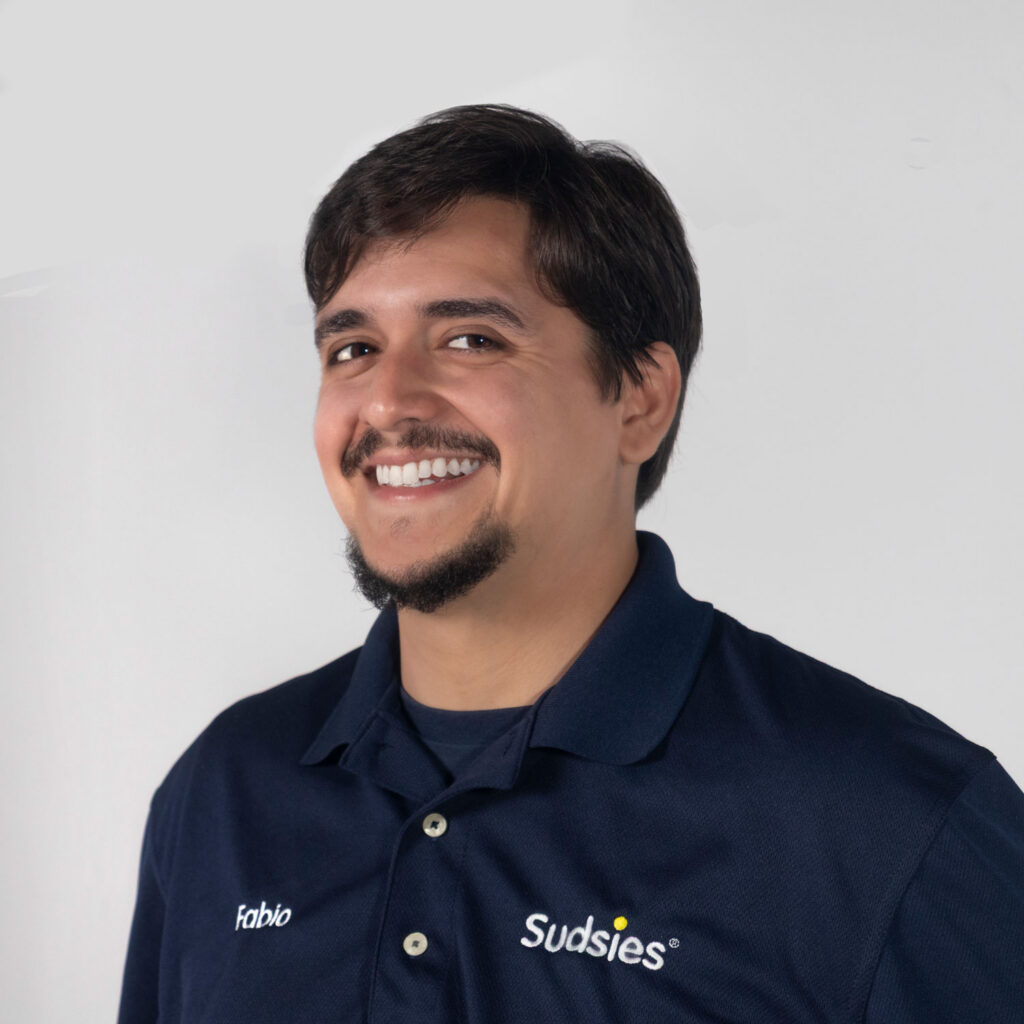 Smiling man wearing a navy polo with 'Fabio' and 'Sudsies' logos, light gray background.