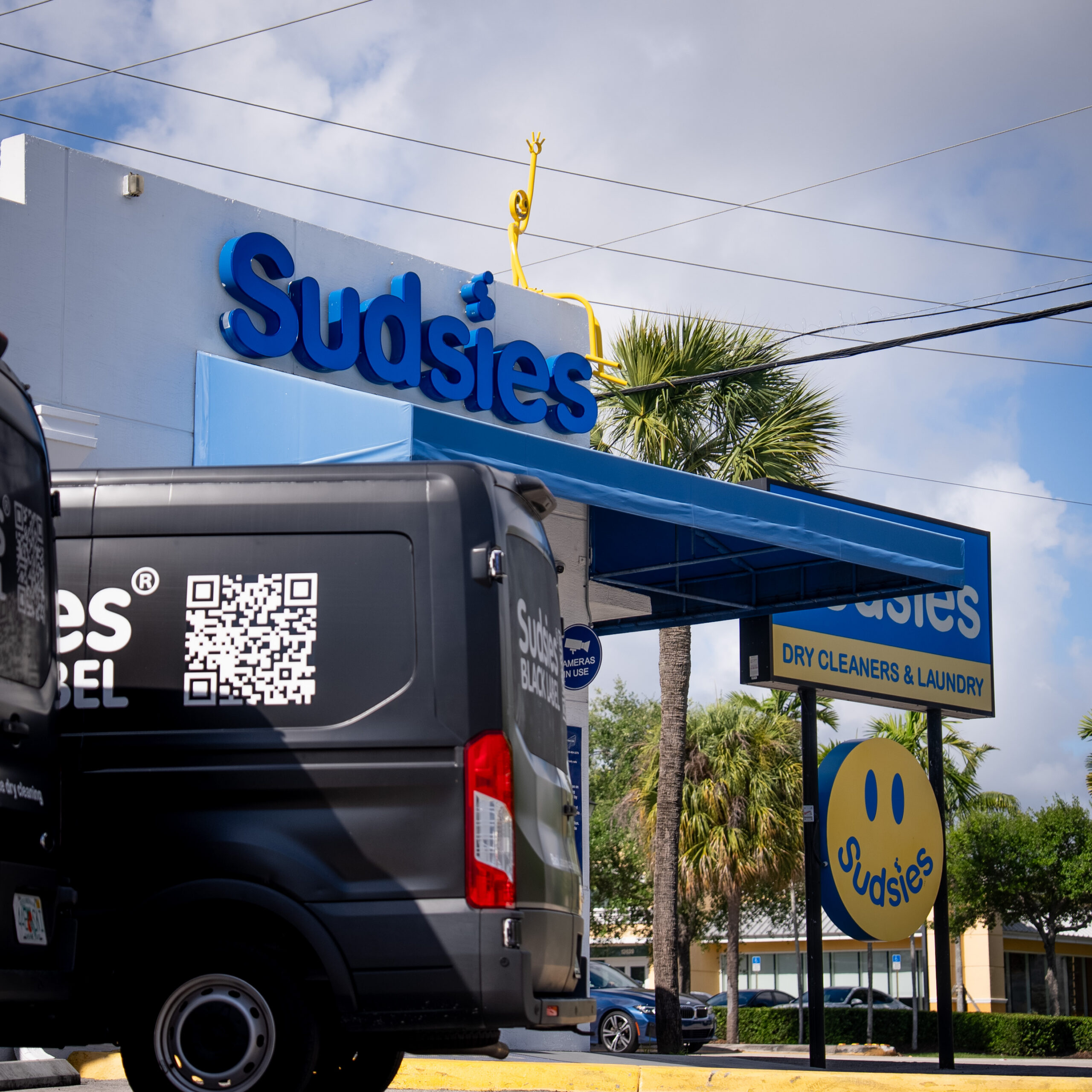 Black van with Sudsies branding and a large QR code on its side, parked near a Sudsies storefront under a blue sky with palm trees in the background.