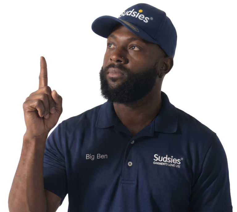 Bearded man in a navy Sudsies polo and cap raises his right index finger, looking upward.