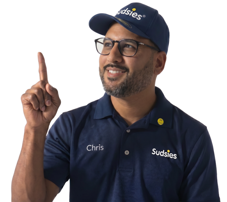 Sudsies garment care valet Chris wearing a navy branded polo, glasses, and cap while pointing upward against a transparent background.