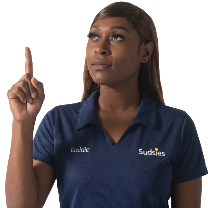 Woman in a navy polo shirt with Goldie and Sudsies logos raises her index finger and looks upward, signaling'one' or drawing attention.