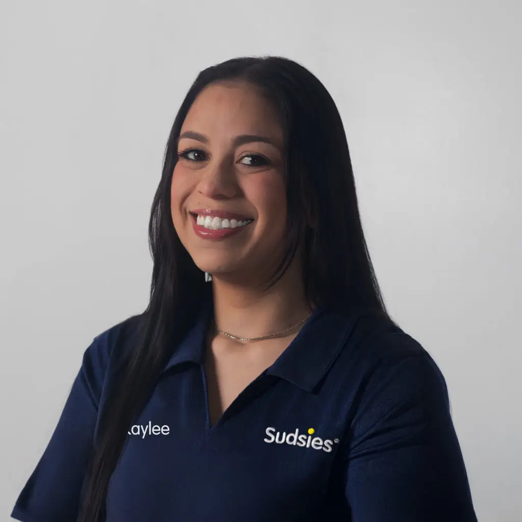 Smiling woman with long dark hair wearing a navy polo with'Kaylee' and 'Sudsies' logos, facing the camera.