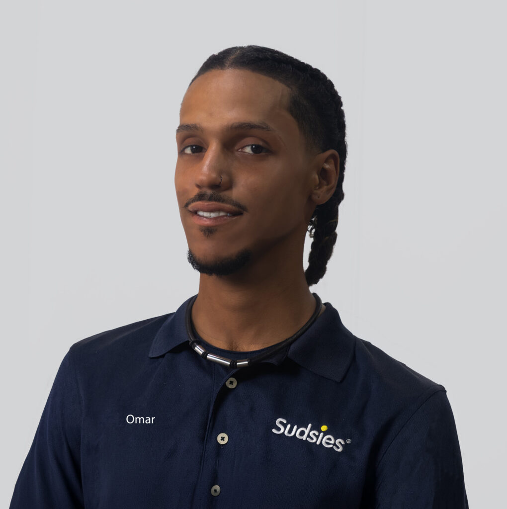Portrait of Omar in a navy polo with Sudsies logo on the chest against a neutral gray background, head-and-shoulders view.