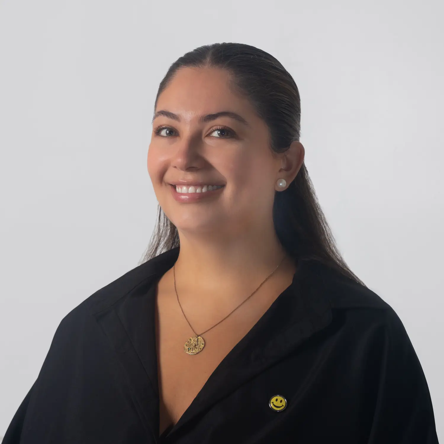 Smiling woman with dark hair in a black blouse, wearing pearl earrings and a gold pendant necklace against a light gray backdrop.
