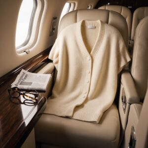 A cream cashmere cardigan draped over a leather seat in a private aircraft cabin, with Financial Times and tortoiseshell glasses on a mahogany table, illustrating designer knitwear