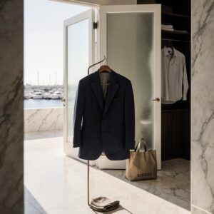 Sunlit marble foyer in Coconut Grove with a worn navy linen blazer on a bronze valet stand, a Sudsies Express garment bag nearby, and a marina view through a salt-sprayed glass doo
