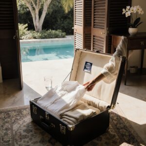 The Seasonal Resident’s Guide to Preparing a Wardrobe Before Leaving South Florida | Sudsies A sunlit marble foyer in a Coconut Grove residence with an open linen trunk, folded garments, and a woman inspecting a shirt cuff for salt spray, illustrating wardrobe care before