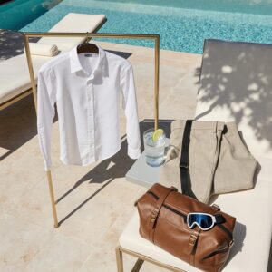 The Snow-to-Sand Guide to Caring for Ski and Resort Wardrobes | Sudsies Open garment bag with dress shirt, cashmere sweater, and linen trousers on a brass stand by a Miami Beach pool, ski goggle strap in leather weekender, quiet luxury editorial scene