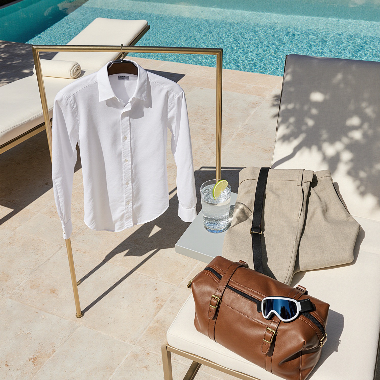 Open garment bag with dress shirt, cashmere sweater, and linen trousers on a brass stand by a Miami Beach pool, ski goggle strap in leather weekender, quiet luxury editorial scene