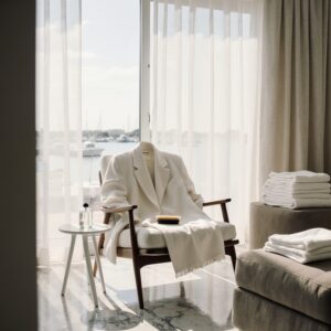 The Vessel Mold Remediation Guide for Textiles | Sudsies | Sudsies Sunlit South Florida living room with marina view, white linen blazer draped on mid-century armchair, brush and cleaning solution on side table, folded bed linens on ottoman, quiet