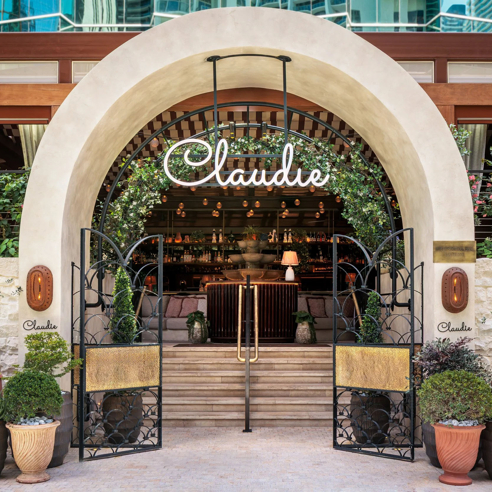 Entrance to a restaurant under a large arch, with greenery, warm lighting, and'Claude' signage on the sides.