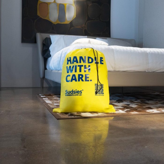 Sudsies yellow laundry bag with “Handle With Care” text placed at the foot of a modern